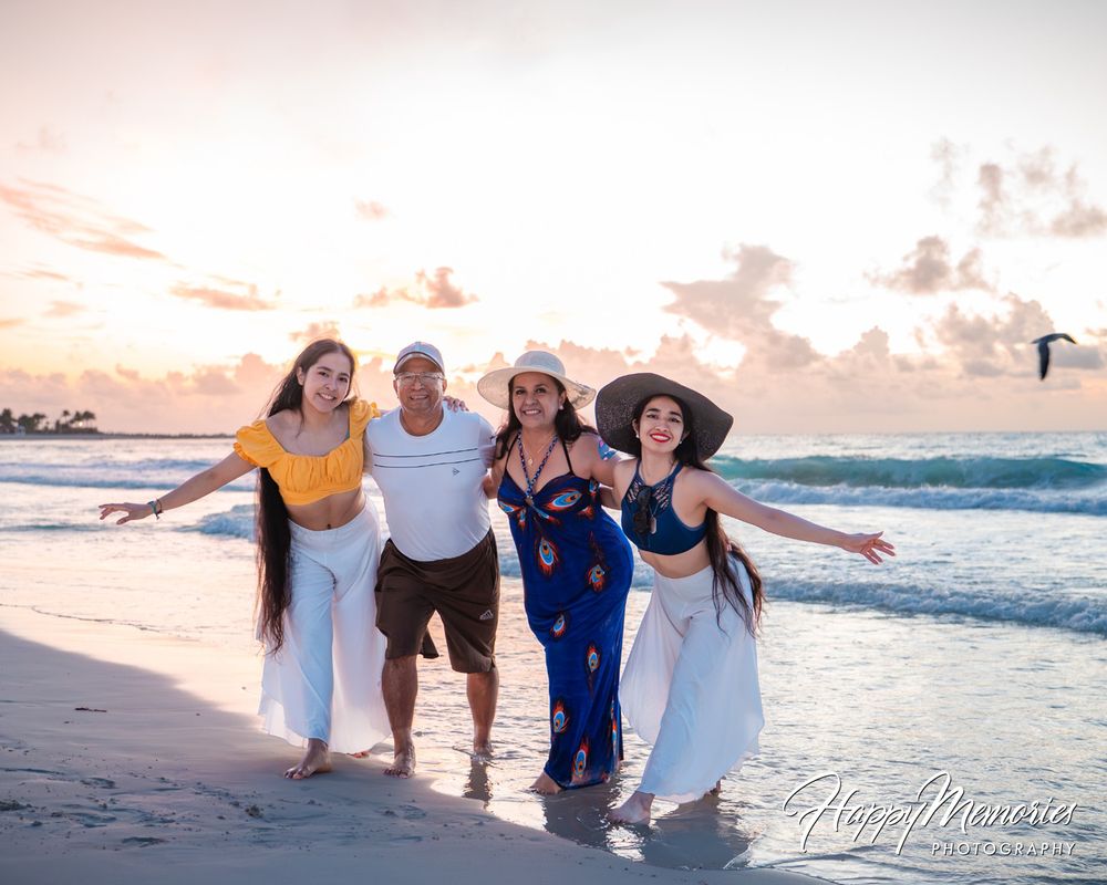 Four smiling family members walk hand in hand along the shoreline at sunset on a sandy beach, with waves and a pink-orange sky behind them.