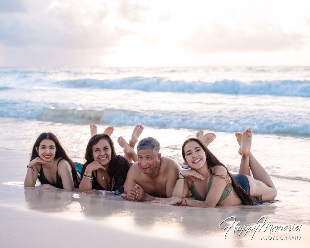 Four people lie side by side on wet sand at the beach, smiling with feet raised as gentle ocean waves roll in at sunset.