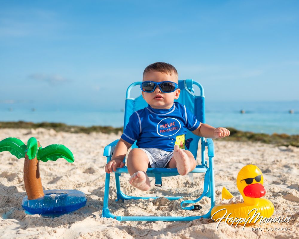 Baby in sunglasses and a blue shirt sits in a blue beach chair on white sand beside an inflatable palm tree and rubber duck.