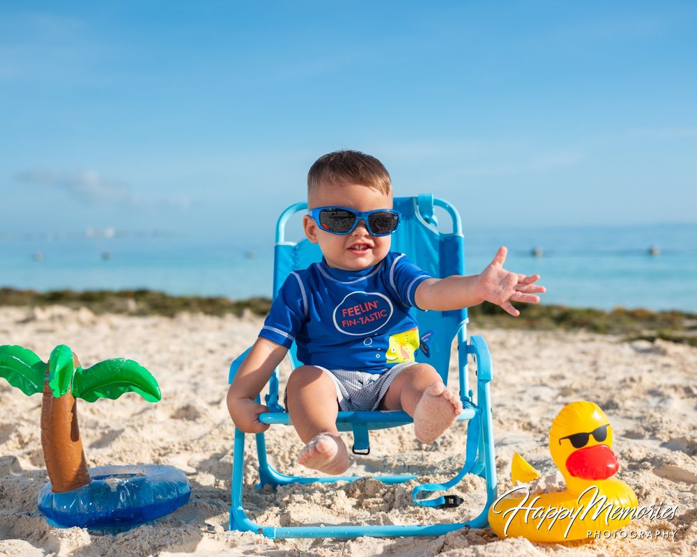Toddler in blue sunglasses and swim shirt sits in a small blue beach chair on sand beside a toy palm tree and rubber duck near the ocean.