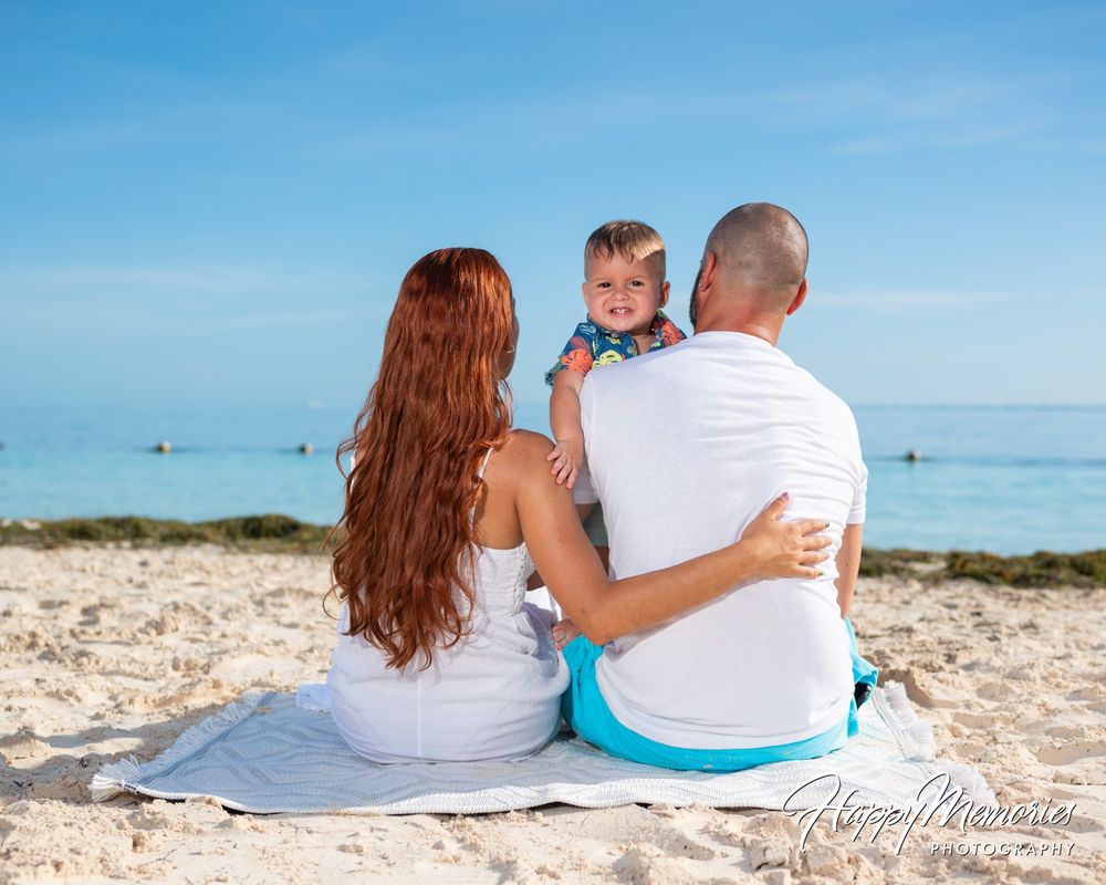 Family of three sitting on a beach blanket facing the turquoise ocean, with a smiling toddler looking back at the camera.