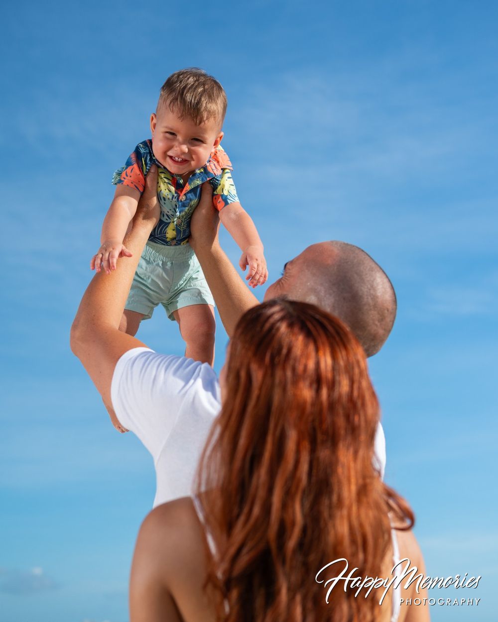Smiling toddler in a colorful shirt and shorts lifted high by a bald man beside a red-haired woman against a bright blue sky.