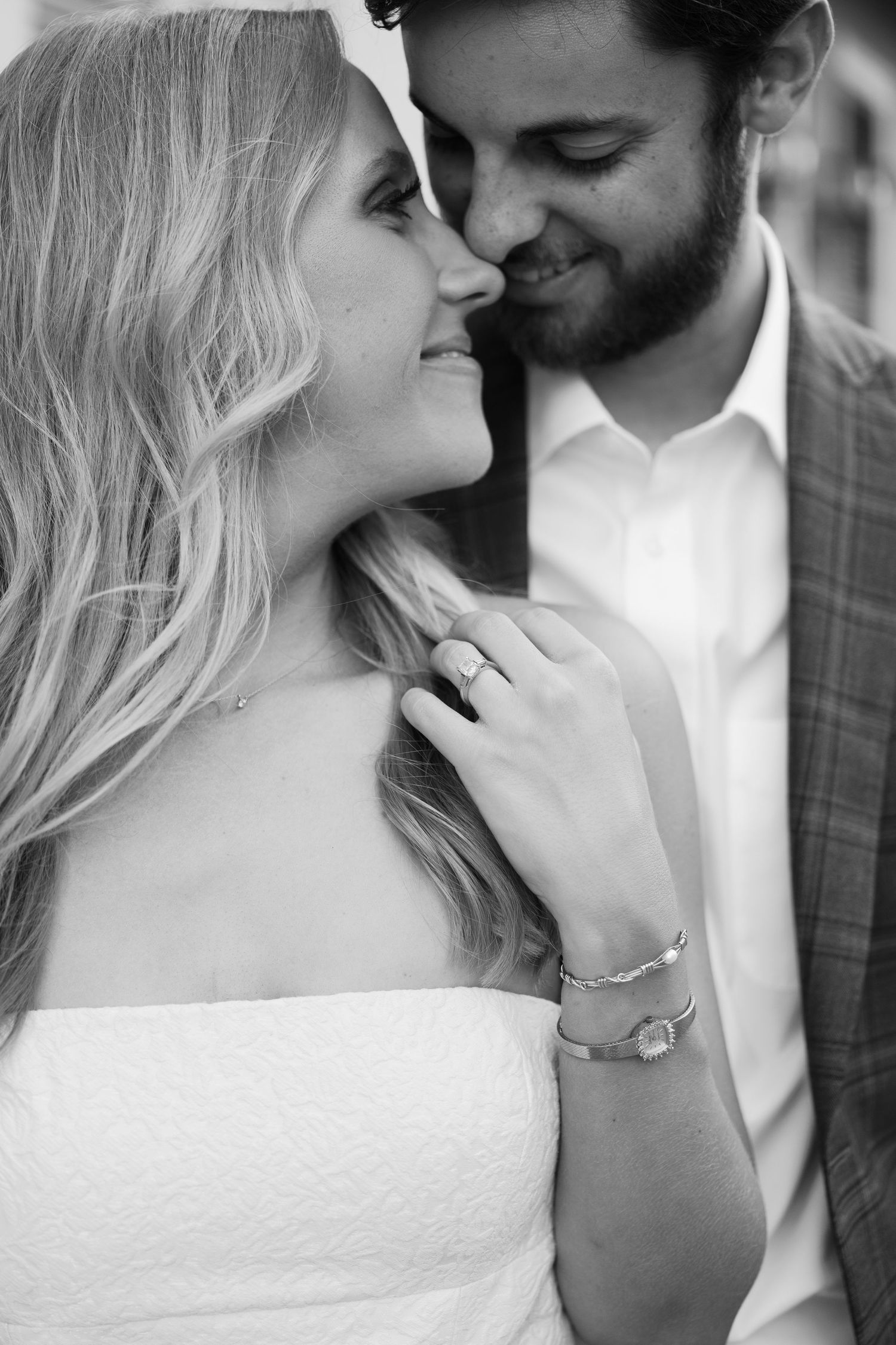 Black and white photo of smiling engaged couple nearly kissing, woman showing diamond engagement ring and bracelet.