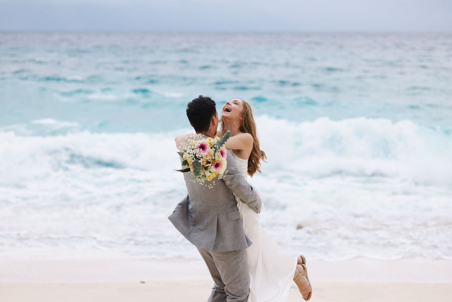 Groom lifts laughing bride holding floral bouquet on beach with crashing waves.