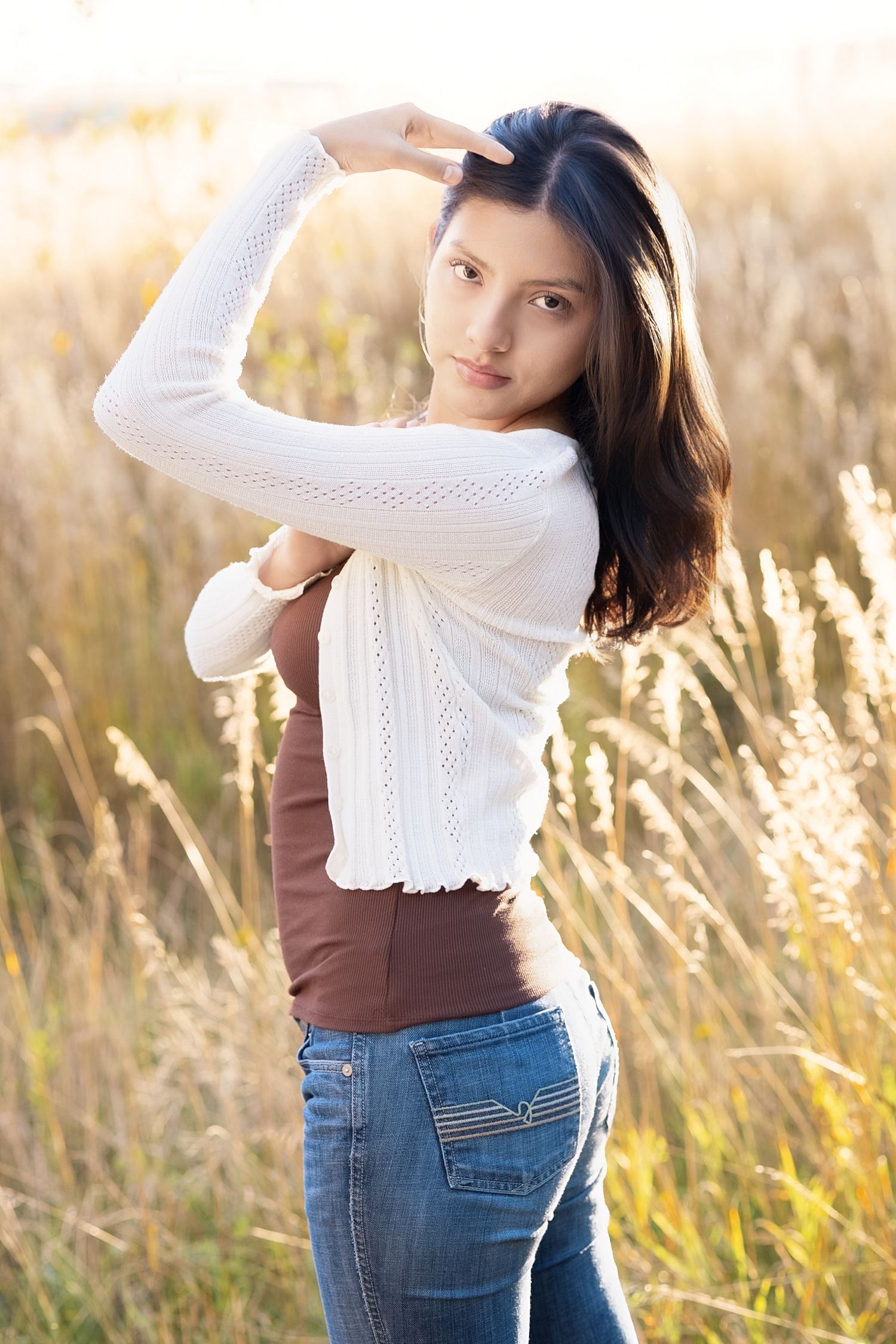 Young woman in white cardigan and jeans poses in golden field, glancing back over shoulder in warm sunlight.