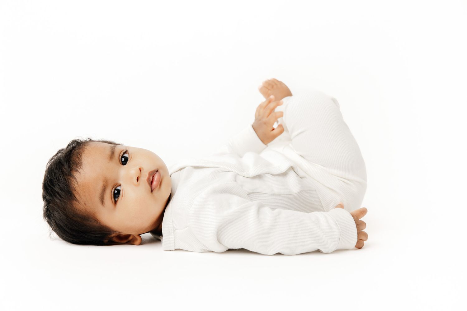 A cute baby in a white onesie sits on a white surface, looking up curiously against a bright white background.