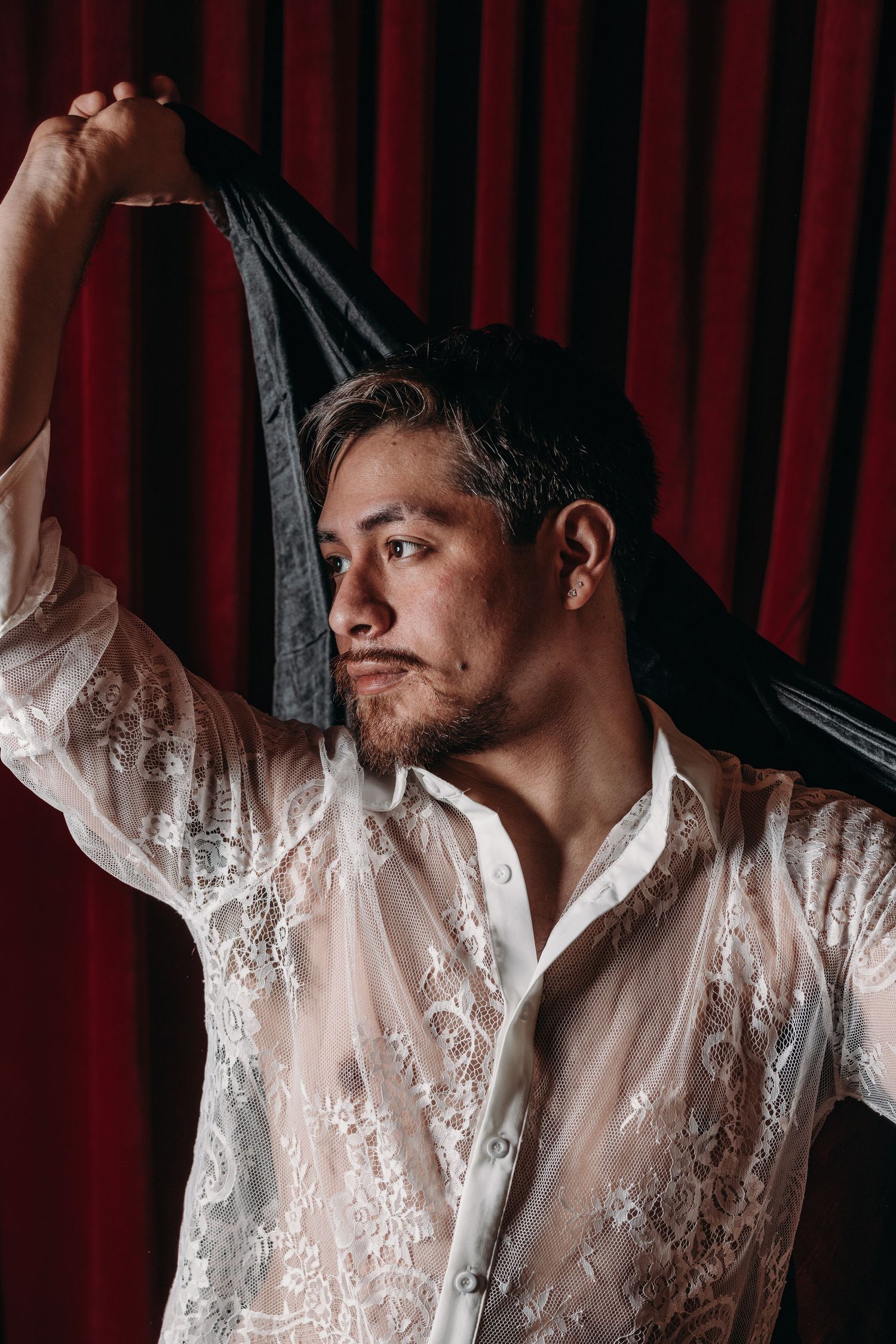 Man with short dark hair and a goatee wearing a sheer white lace shirt raises a black sheer cloth overhead against a red velvet curtain.