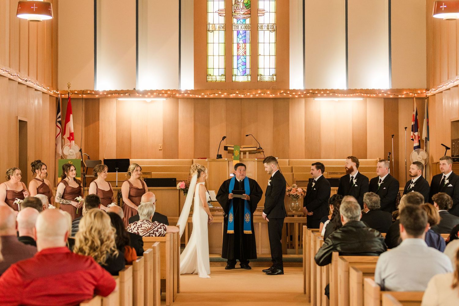 Wedding ceremony in a warmly lit church with bridesmaids, groomsmen, and officiant at the altar.