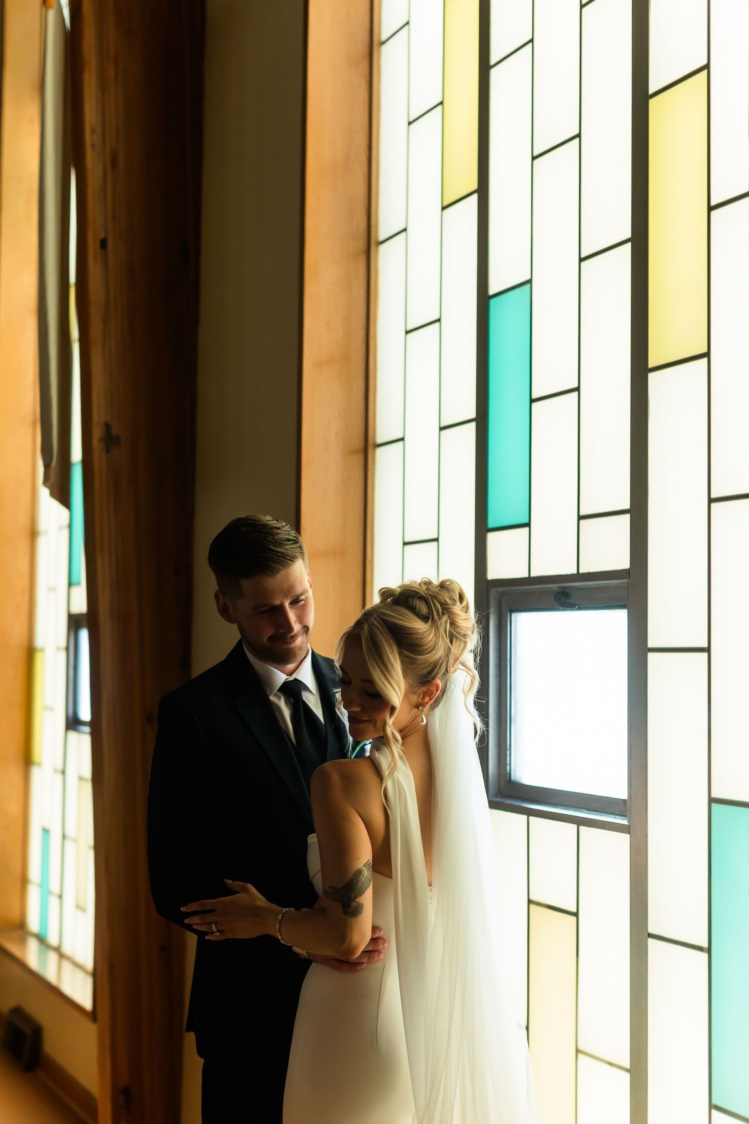 Bride and groom embrace before stained glass windows, bride in white gown with veil, groom in black suit.