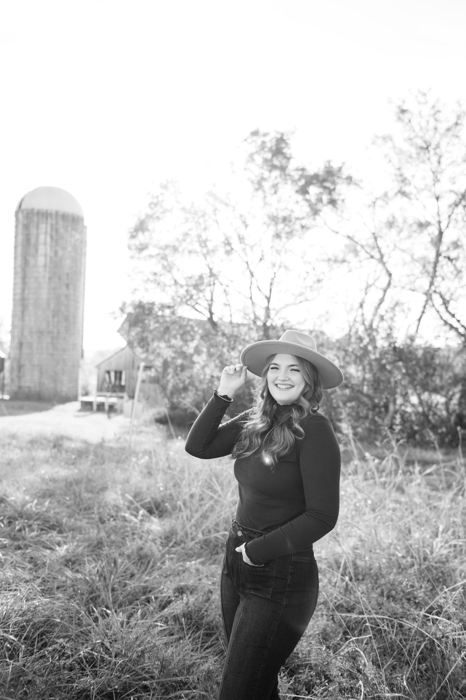 Senior girl  in Black and white photo of a smiling young woman in a wide-brim hat and black outfit posing near a farm silo.