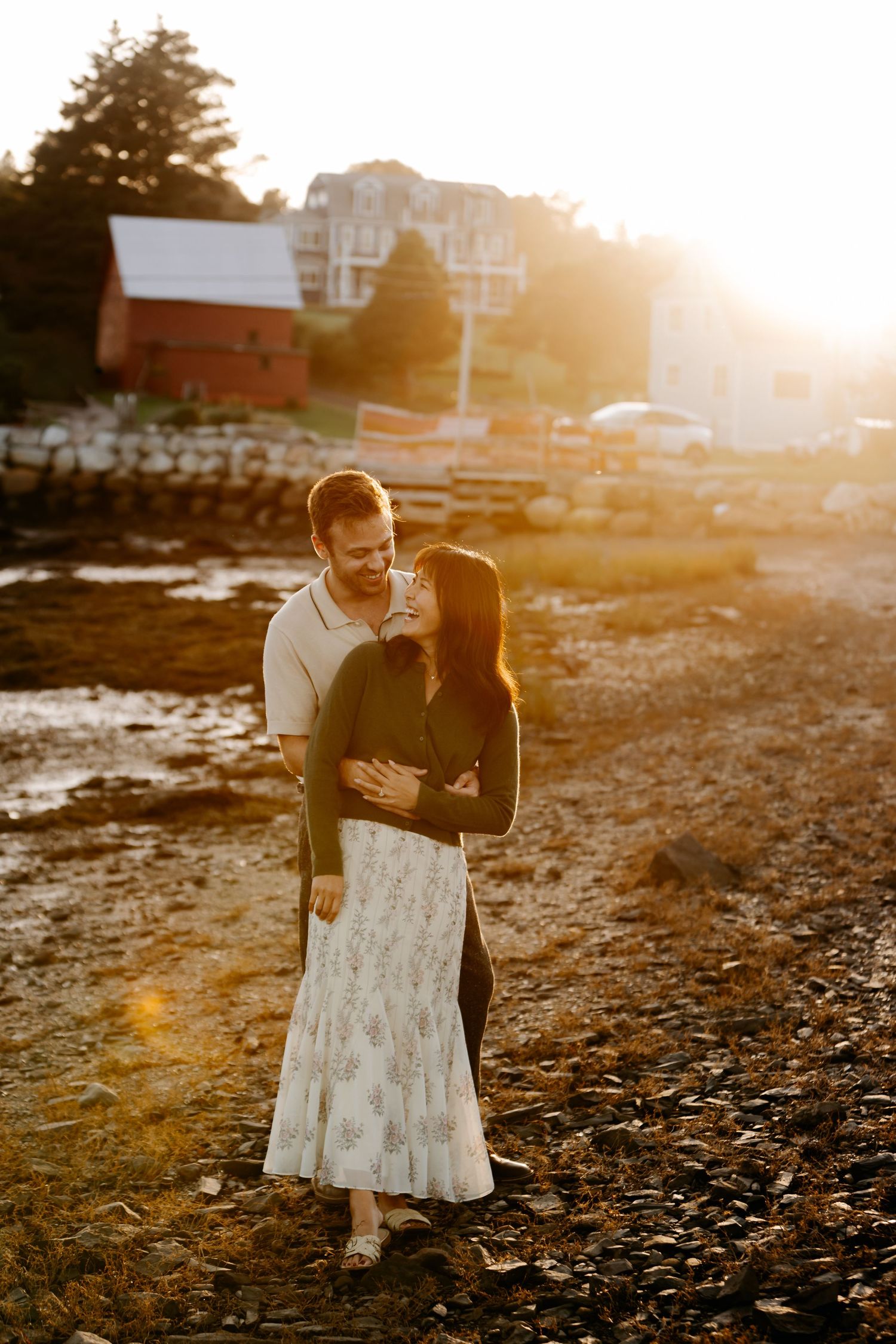 Couple embracing at golden hour on a rocky shoreline with a red barn and waterfront buildings in the background.