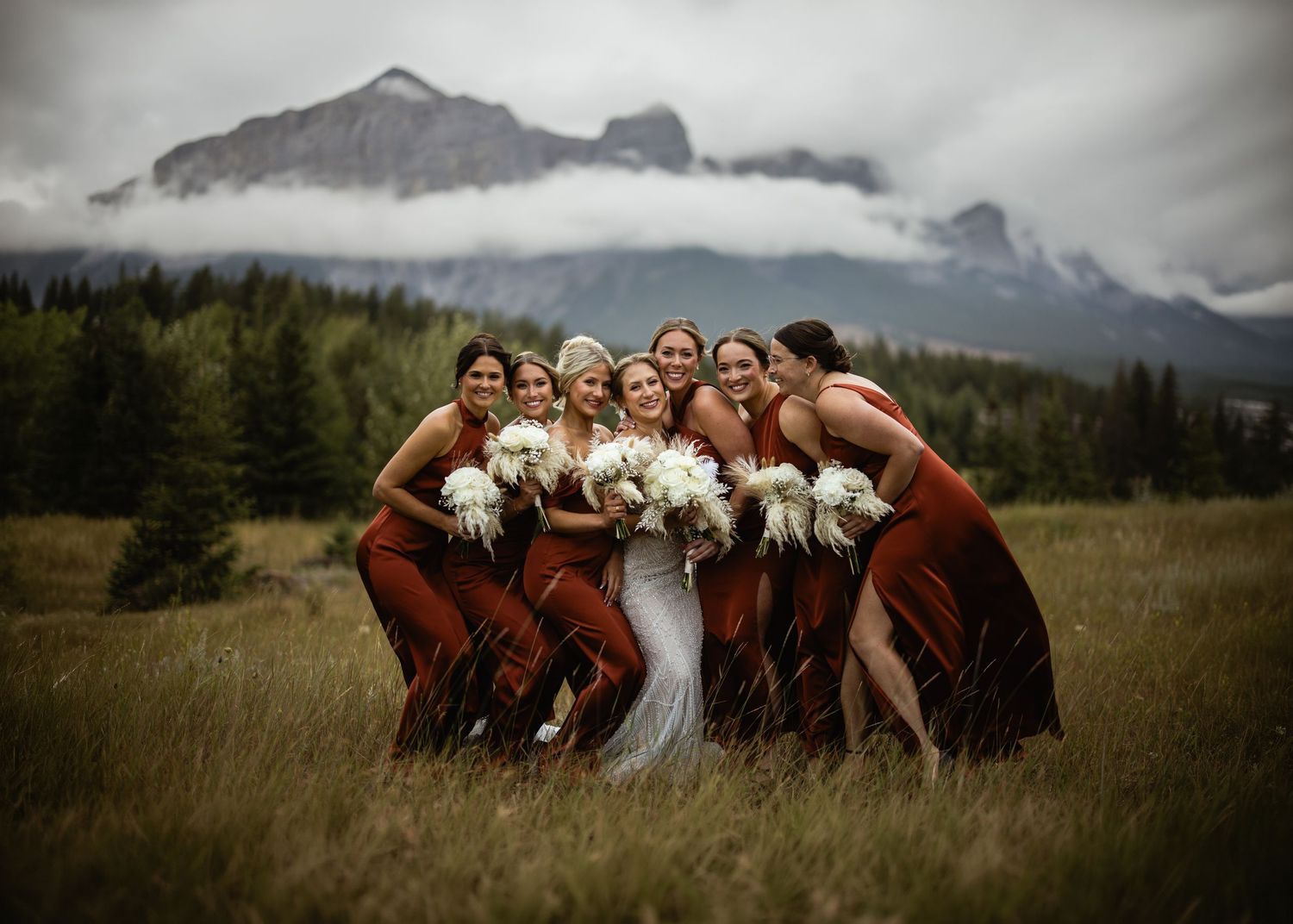 Laughing bride and bridesmaids in rust gowns in a mountain meadow under low clouds, by a Canmore wedding photographer