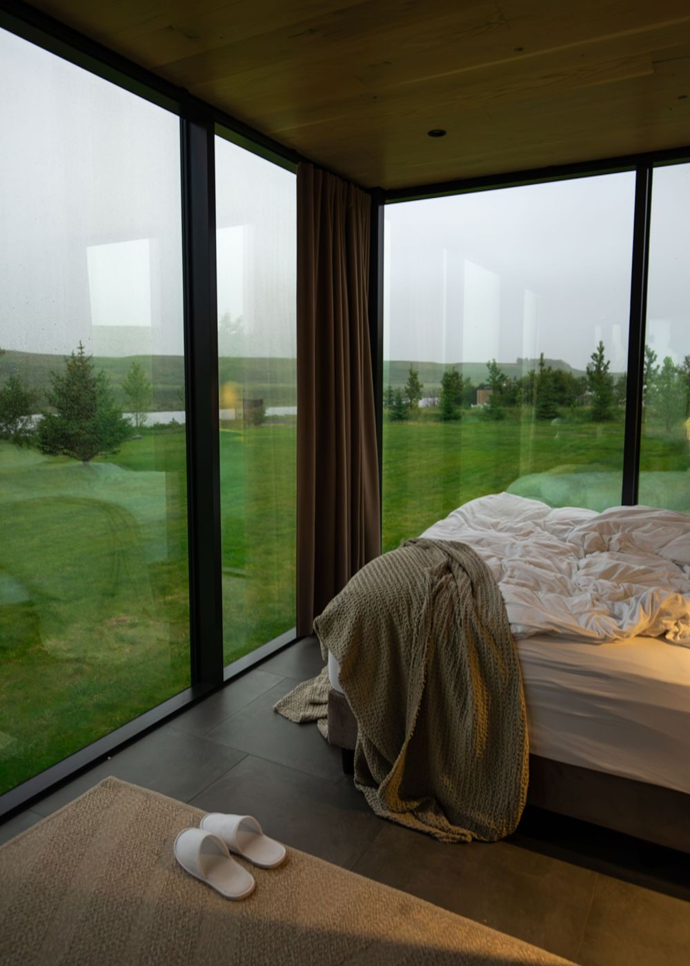 Minimalist bedroom with floor-to-ceiling windows overlooking a misty green landscape, featuring a cozy bed and white slippers.