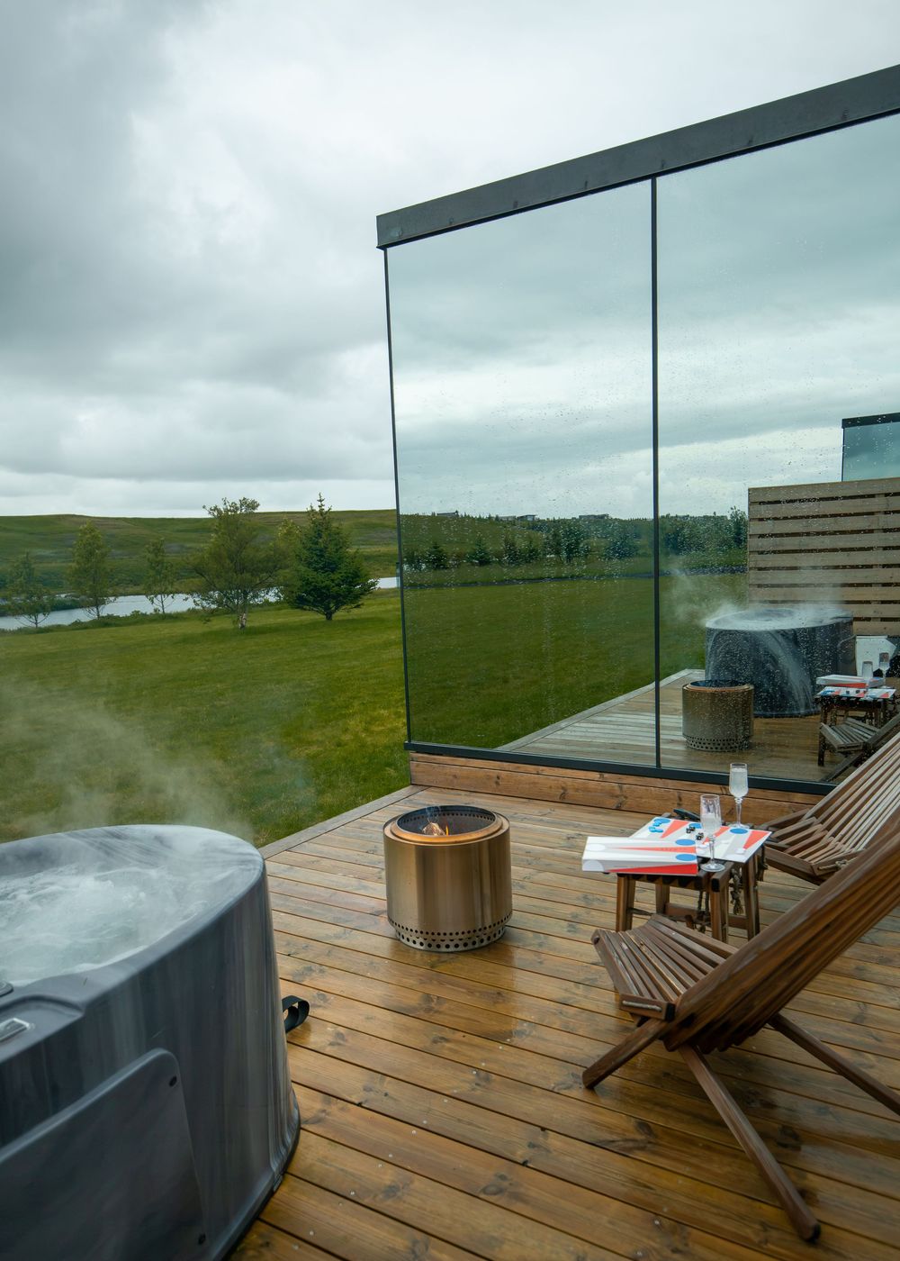 Steaming hot tub on a wooden deck with glass walls, lounge chair, fire pit, and scenic green landscape views.