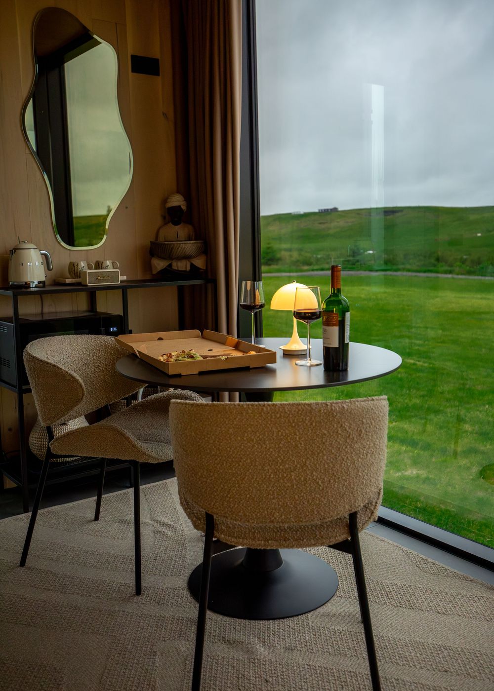 Cozy hotel room dining area with pizza, wine, and a glowing lamp beside a large window overlooking green hills.