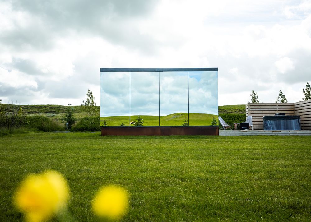 Mirror-clad modern building reflecting green landscape under cloudy sky, surrounded by lush grass with yellow flowers in foreground.