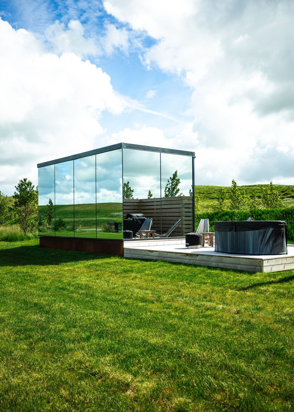 Modern glass cube cabin with wooden deck, hot tub, and lush green lawn under a partly cloudy sky.