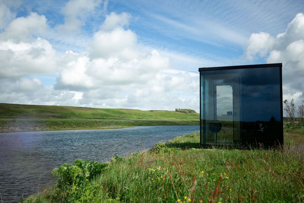 Modern glass structure beside a serene river with lush green hills and dramatic cloudy sky in rural landscape.