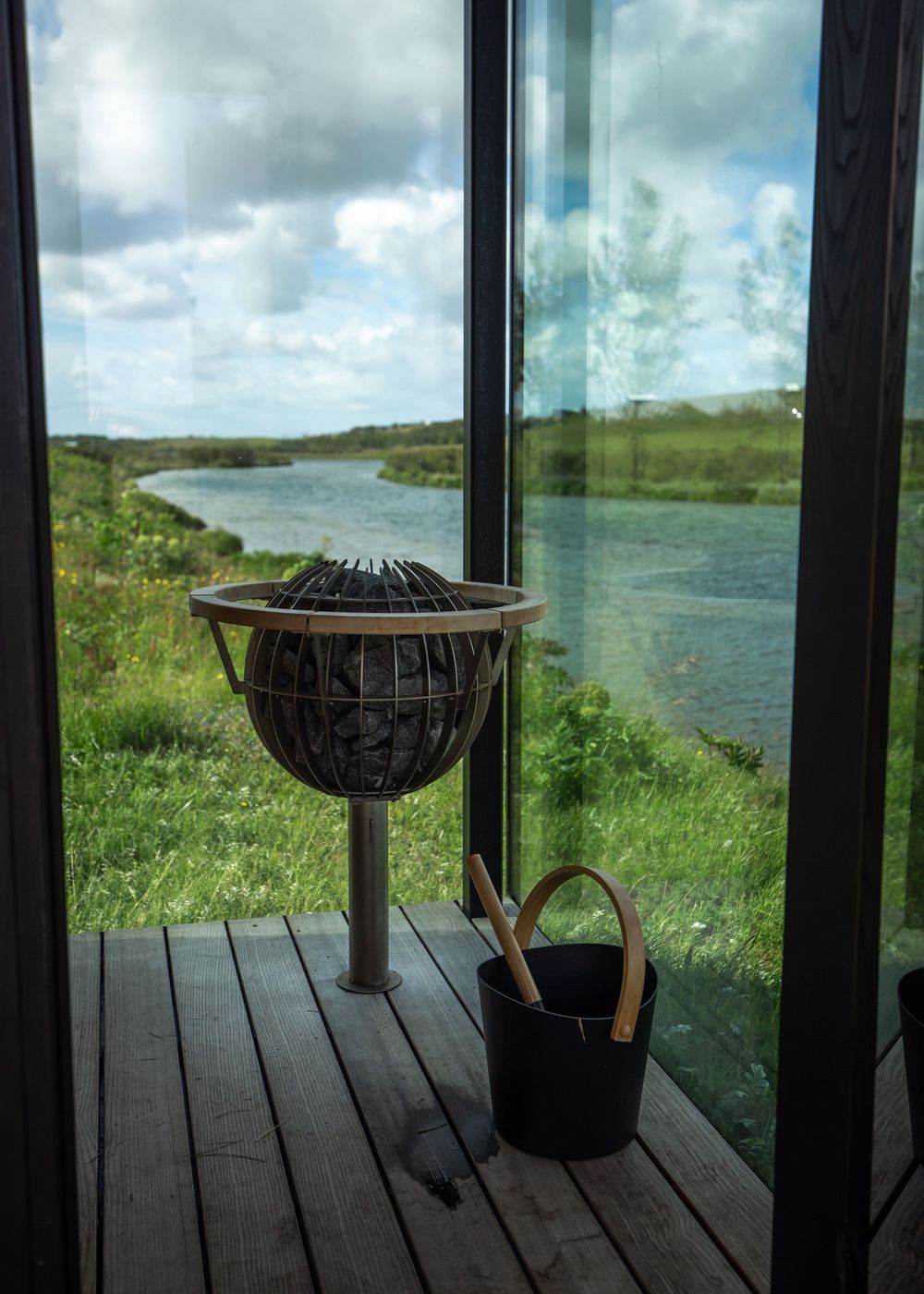 Sauna stove with bucket and ladle on wooden deck overlooking scenic river and green landscape through glass windows.