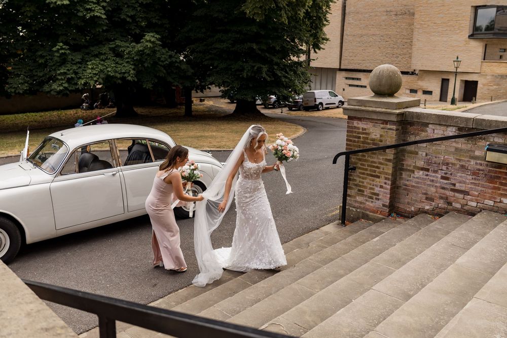 Bride in white gown assisted by bridesmaid on steps beside a classic white wedding car.