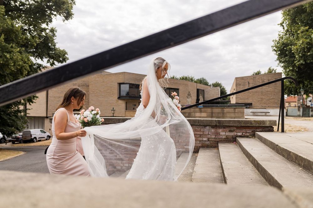 Bride in white gown with flowing veil walks up steps assisted by bridesmaid in pink dress outdoors.