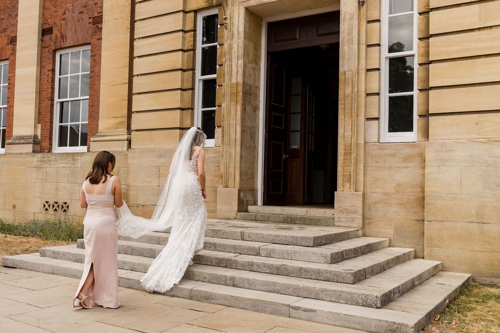 Bride in white lace gown and veil walks up stone steps with bridesmaid in pink dress at elegant venue.