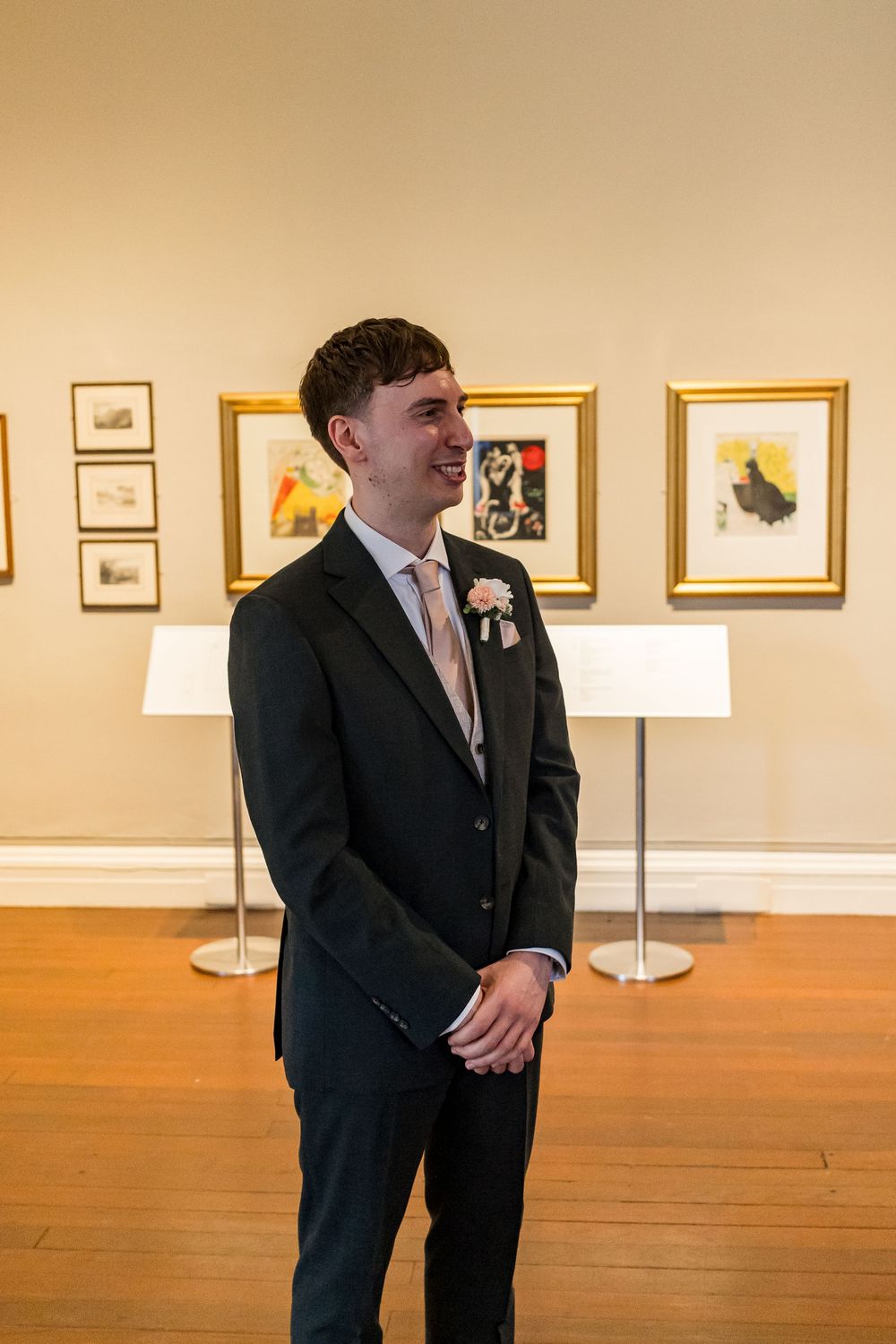 Smiling groom in dark suit with floral boutonniere stands in art gallery before wedding ceremony.