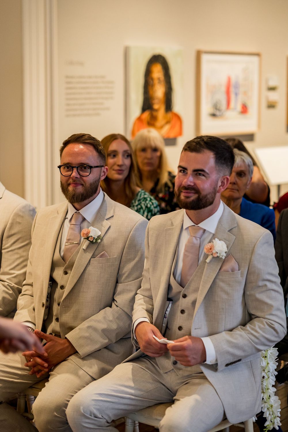 Two groomsmen in matching beige suits with floral buttonholes smile during a wedding ceremony in an art gallery.