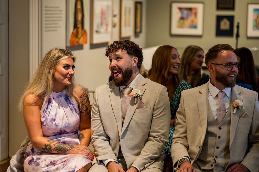 Joyful wedding guests laughing during ceremony, groom in tan suit with boutonniere seated beside smiling blonde woman.