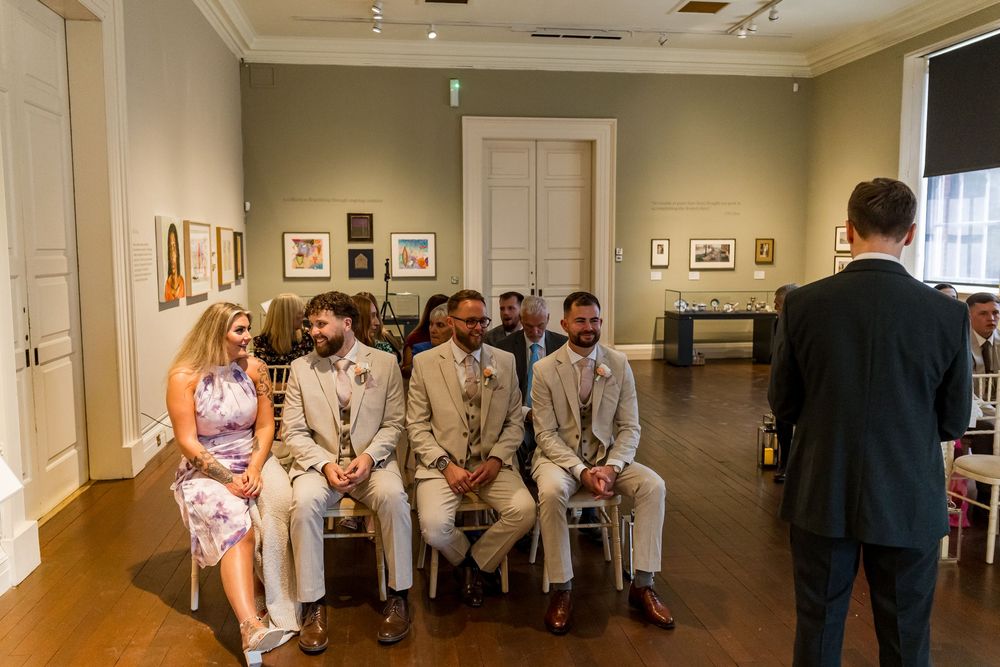 Wedding guests seated in an art gallery ceremony venue, smiling at the officiant during an indoor wedding.