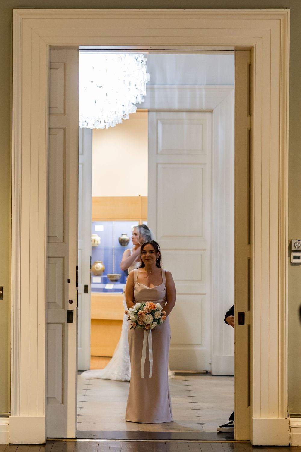 Bridesmaid in champagne gown holds floral bouquet standing in elegant doorway with bride visible behind her.
