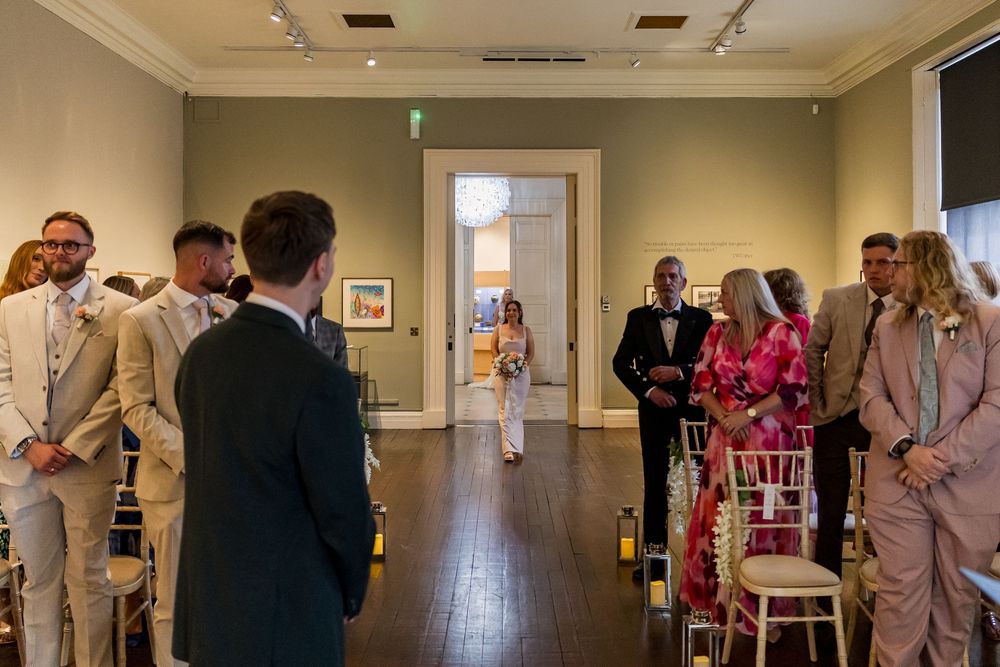 Bride walks down the aisle escorted by her father in an elegant art gallery wedding ceremony venue.
