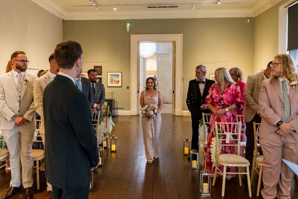 Bride walking down the aisle in a white pantsuit at an elegant art gallery wedding ceremony.