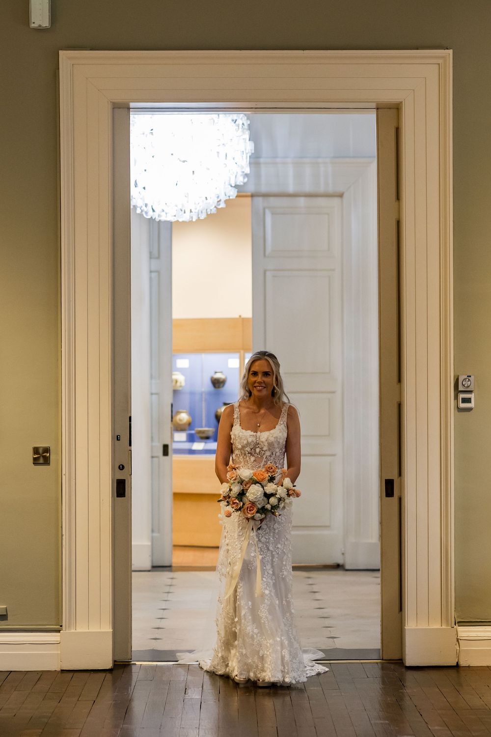 Smiling bride in a fitted lace wedding gown holding a bouquet, standing in an elegant doorway with a crystal chandelier.