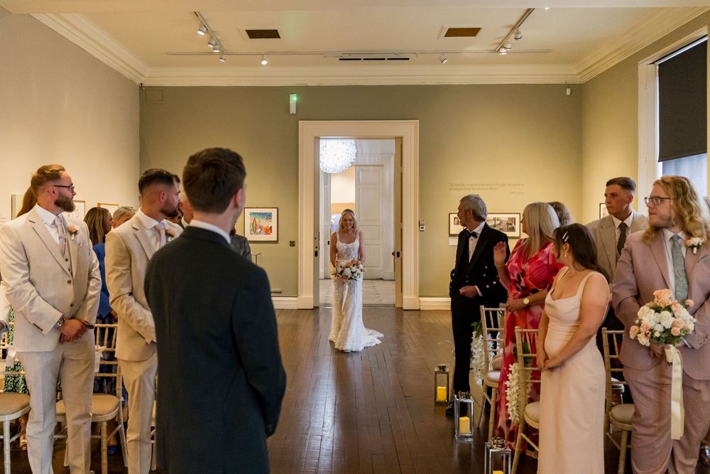 Bride walks down aisle escorted by family in elegant art gallery wedding ceremony venue.