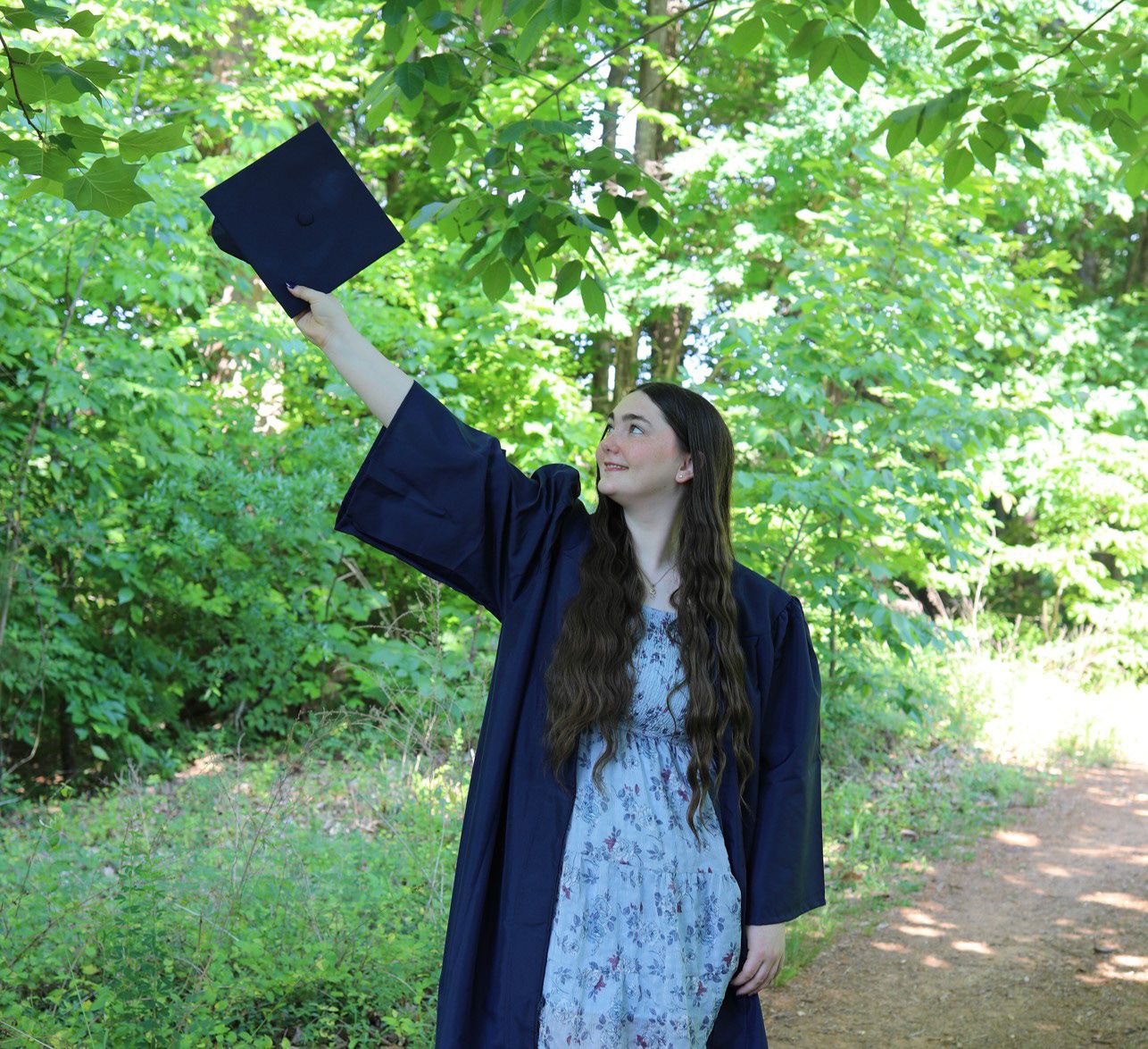 Young woman in navy graduation gown raises cap triumphantly on a sunny forest path, celebrating her academic achievement.