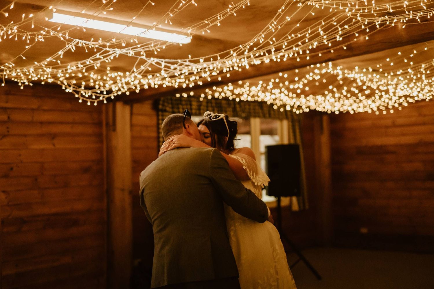 Couple sharing first dance under warm fairy lights in a rustic wooden barn at their wedding reception.