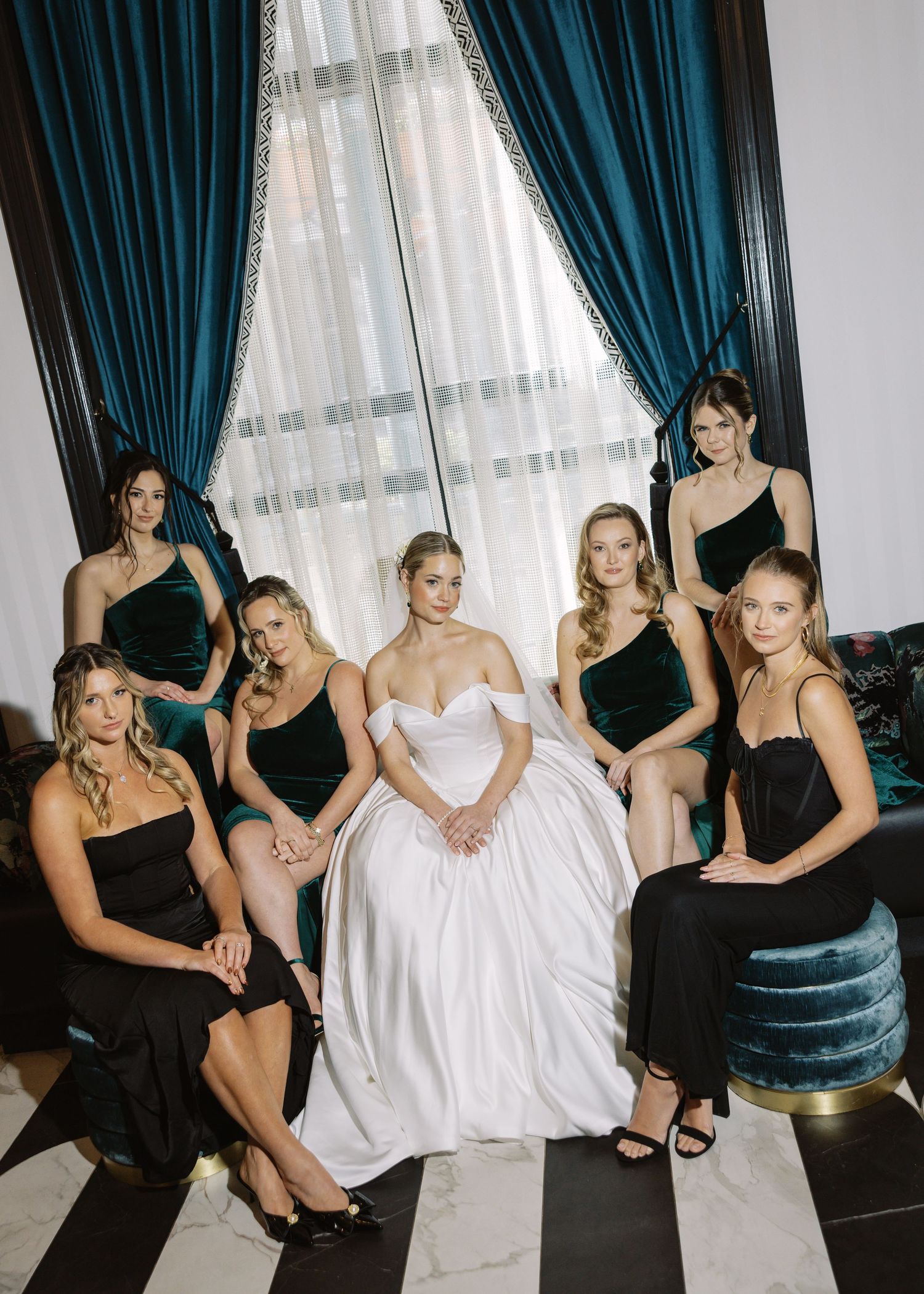 Bride in white gown poses with six bridesmaids in black dresses near teal curtains in an elegant room.