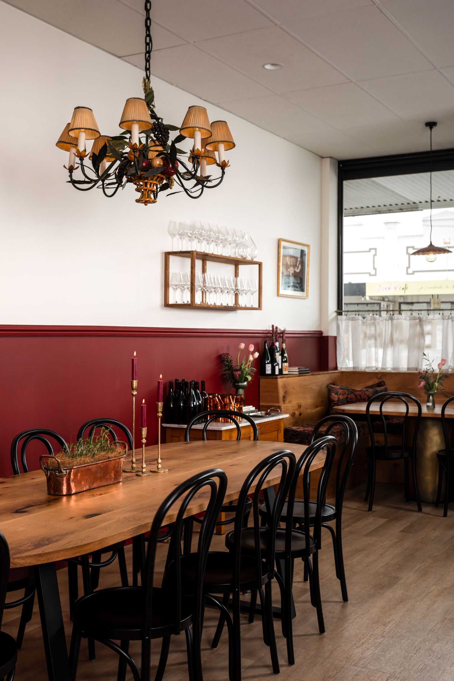 Cozy restaurant interior with a long wooden table, black bentwood chairs, burgundy wainscoting, candles, and a chandelier with amber shades.