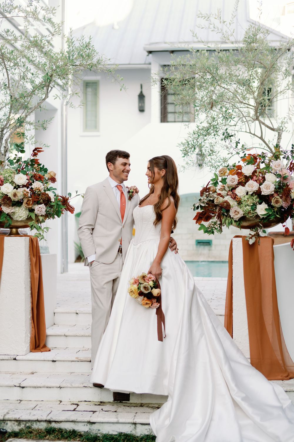 Bride and groom smiling together outdoors with lush floral arrangements and pool backdrop.