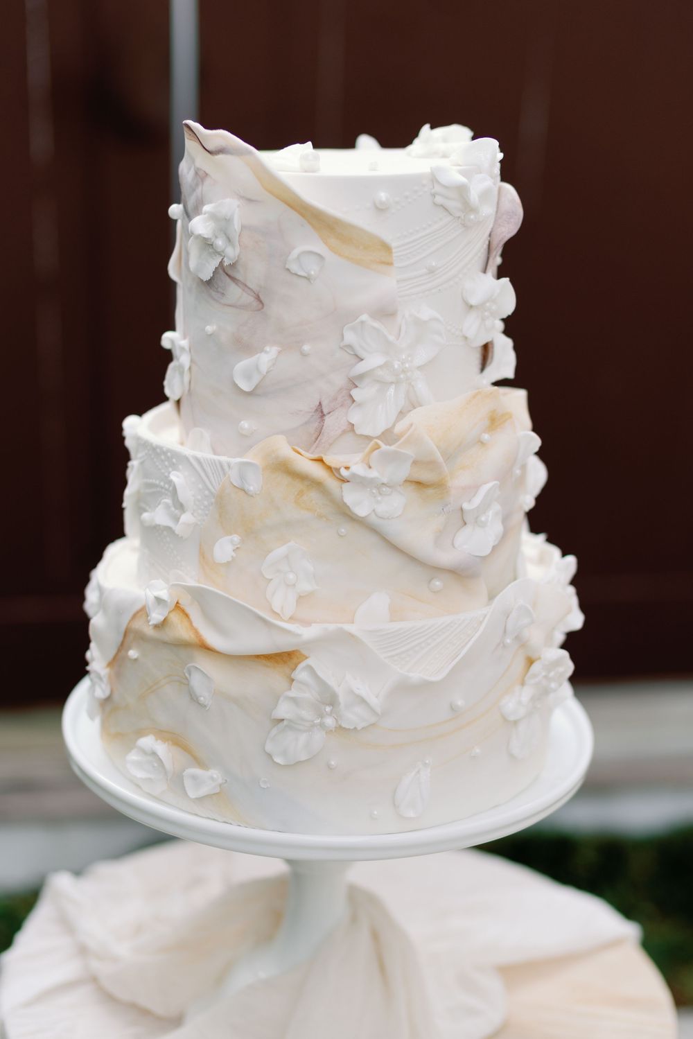 Three-tier white wedding cake with floral appliques and draped fondant details on cake stand.