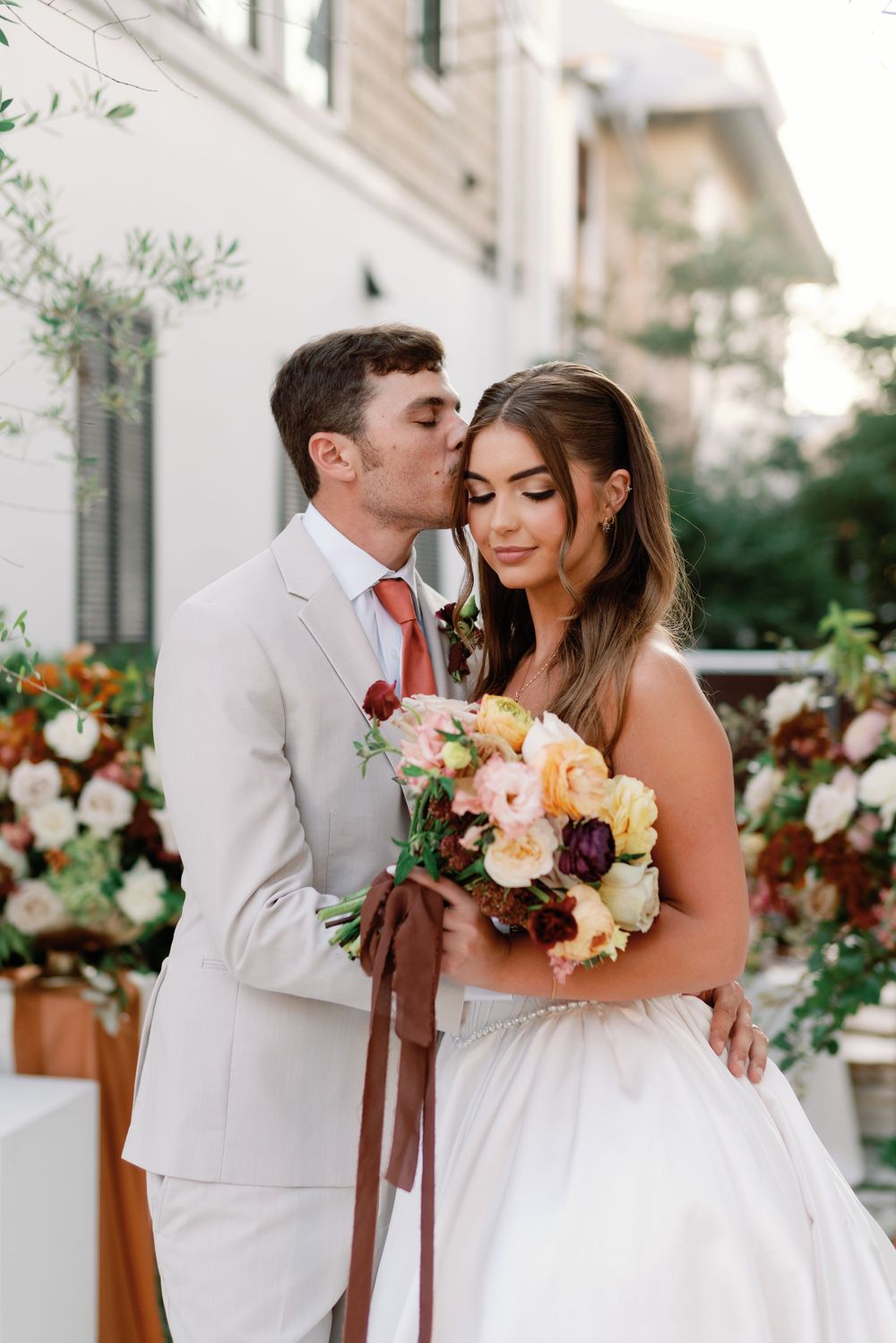 Groom kissing bride on cheek as she holds colorful bouquet with ribbon at outdoor wedding.