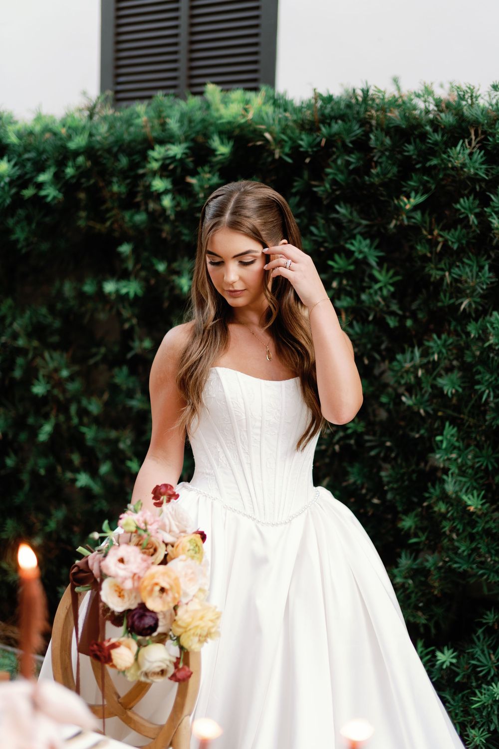 Bride in strapless corset ballgown holds colorful bouquet of roses and ranunculus against lush green hedge backdrop.