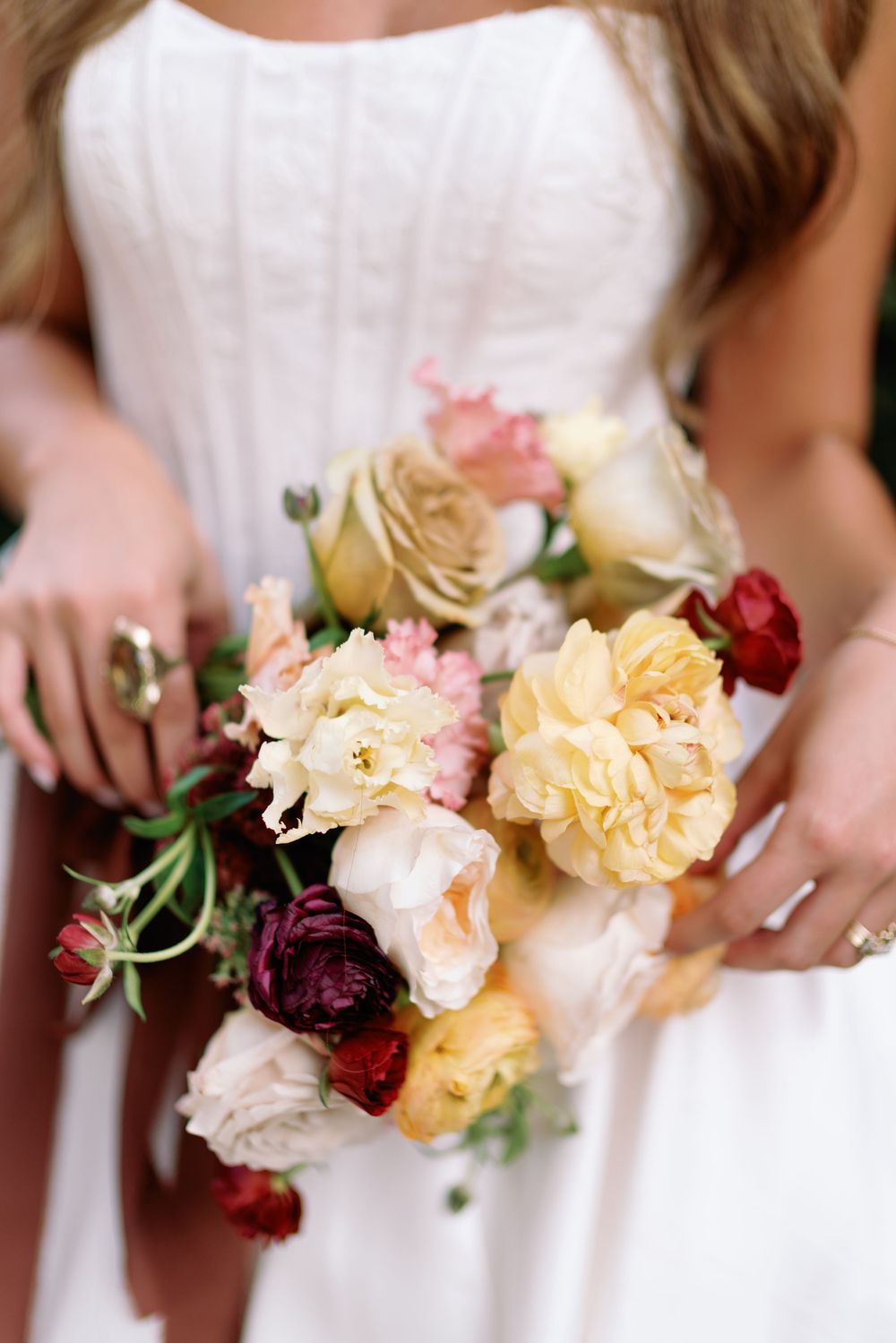 Close-up of bridal bouquet featuring cream, blush, burgundy, and yellow roses and ranunculus held by bride in white gown.