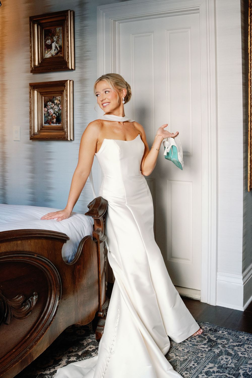 Smiling bride in sleek strapless white gown holding teal shoes beside antique wooden bed.