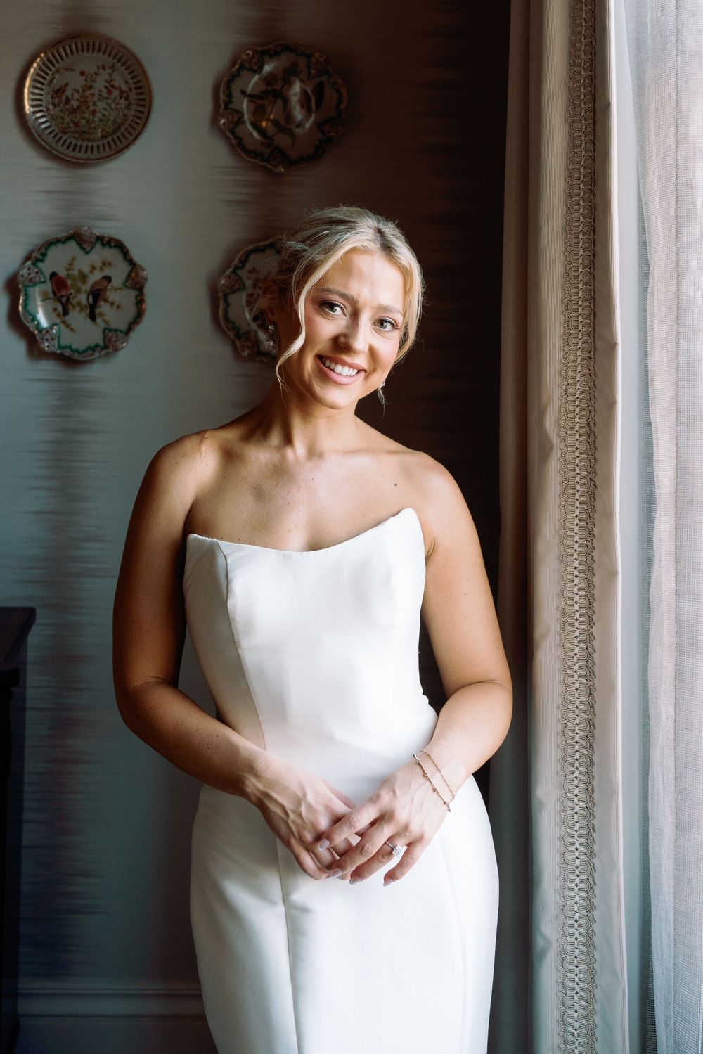 Radiant bride in minimalist strapless white gown smiling by window with decorative plates.