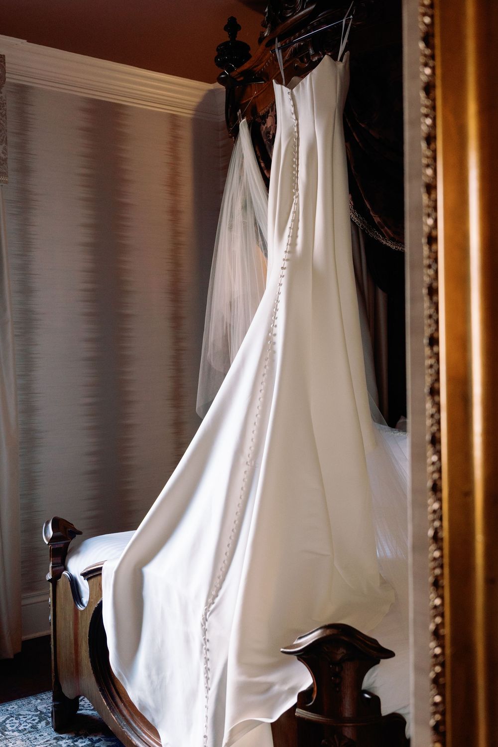 Elegant white wedding gown with button back and veil hanging on ornate dark wood four-poster bed.