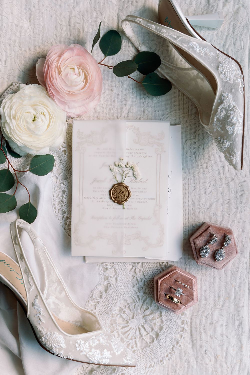 Bridal flat lay featuring lace heels, pink velvet ring box, floral invitation with wax seal, and roses.