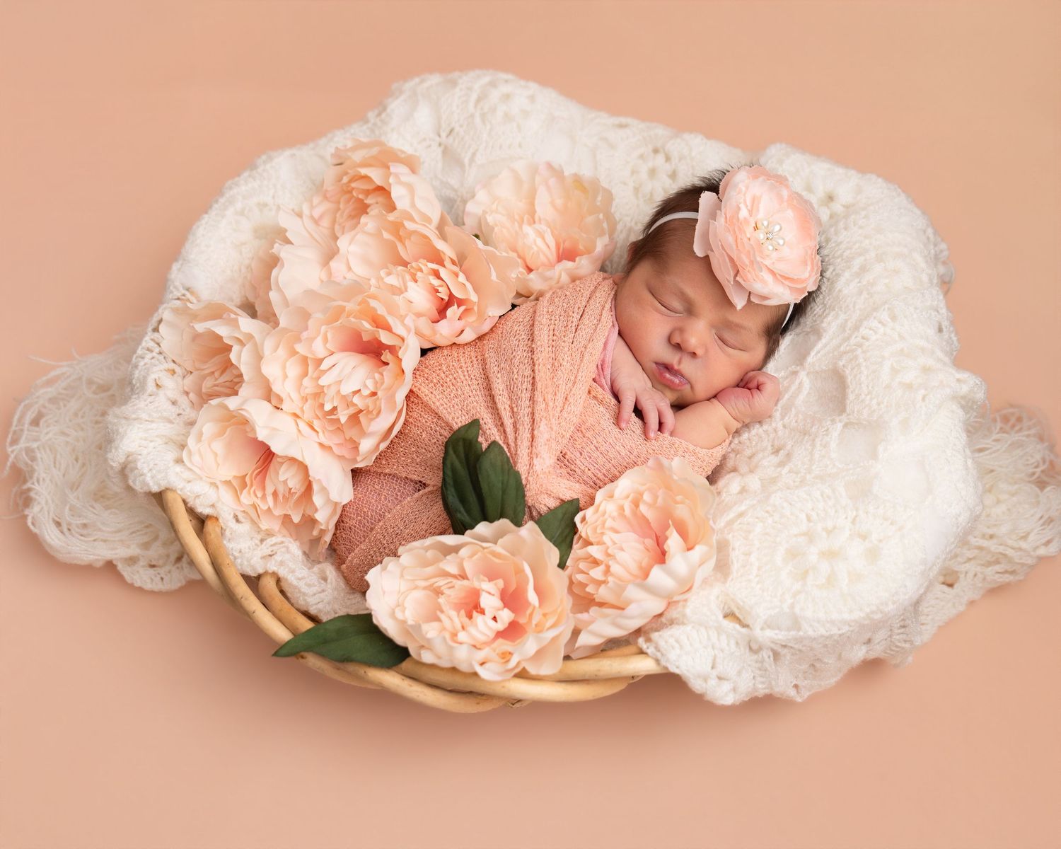 Sleeping newborn baby wrapped in peach swaddle with floral headband, nestled in wicker basket with lace and peach flowers.