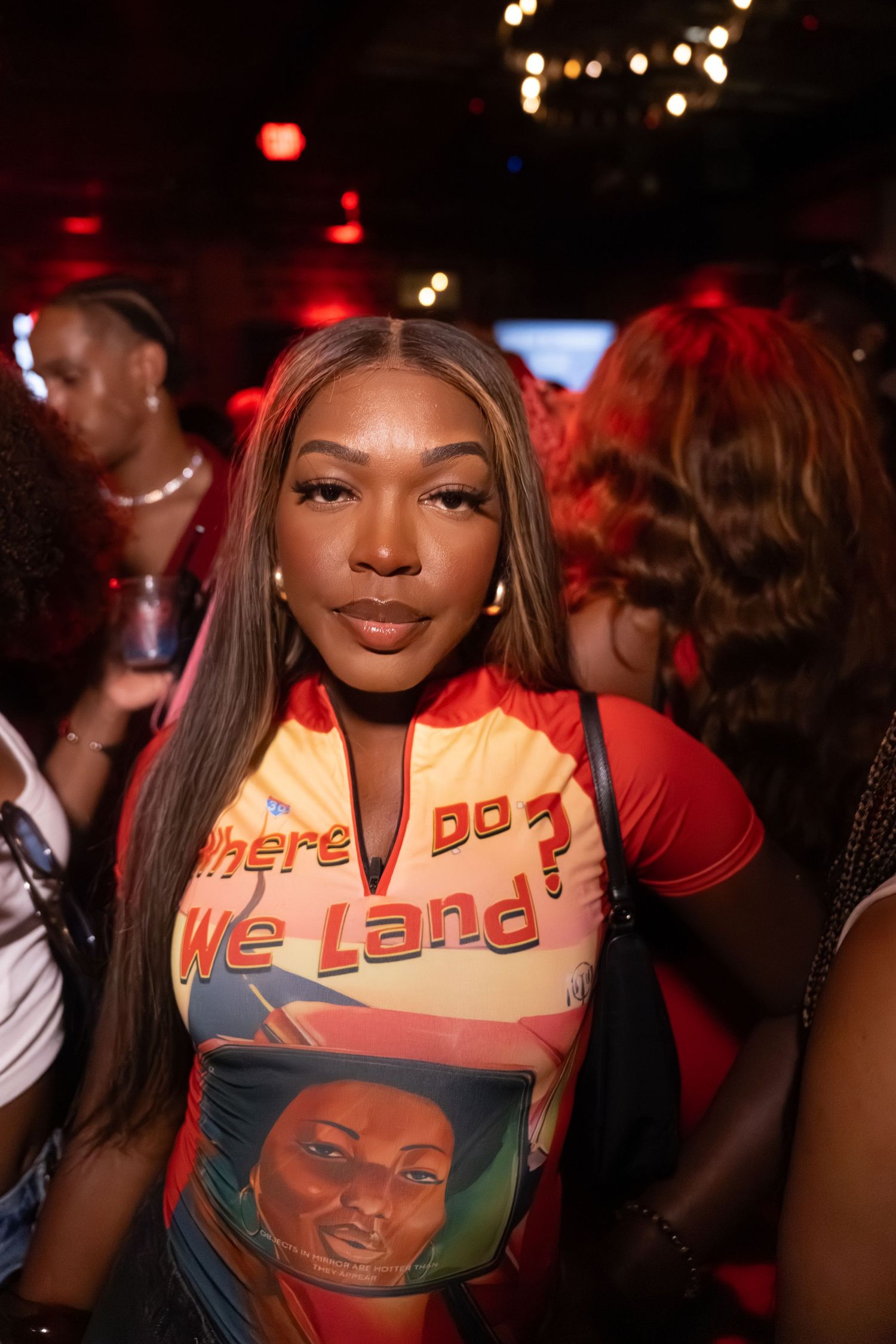 Young woman in colorful graphic tee reading 'Where Do We Land?' poses at a dimly lit nightclub event.
